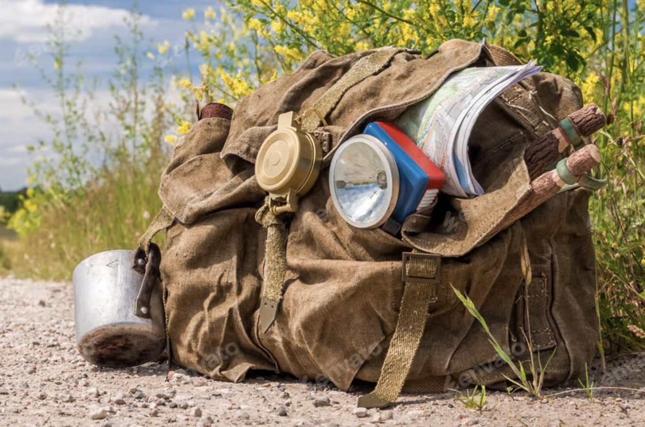 A heavily overloaded vintage canvas rucksack on a gravel roadside, hung with a compass, torch, map and cooking pot