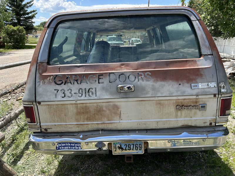 Scott's rusted GMC Suburban hand-lettered “Scott Garage Doors”, with a bumper sticker joking that you have to be secure to be seen in a car like this