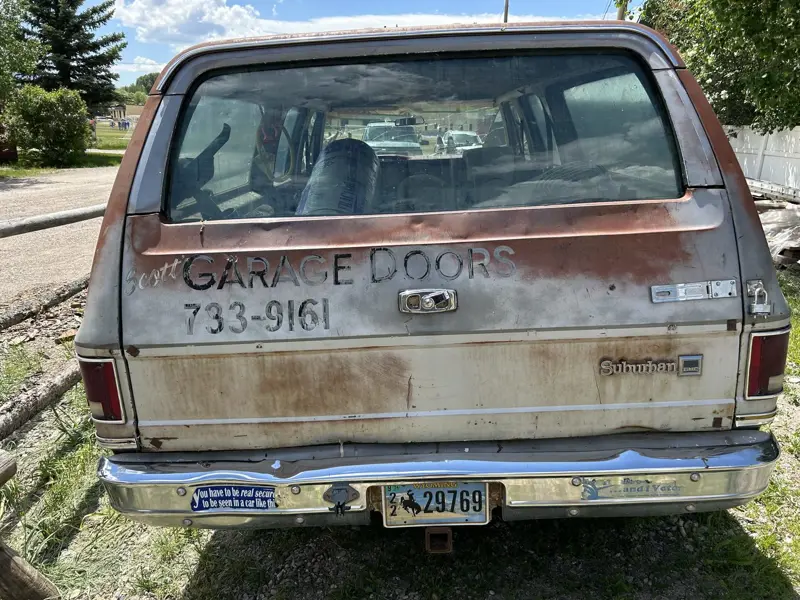 Scott's rusted GMC Suburban hand-lettered “Scott Garage Doors”, with a bumper sticker joking that you have to be secure to be seen in a car like this