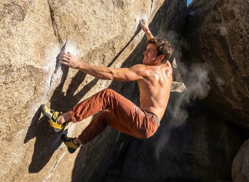 A shirtless climber bouldering across a steep rock overhang, chalk dust in the air