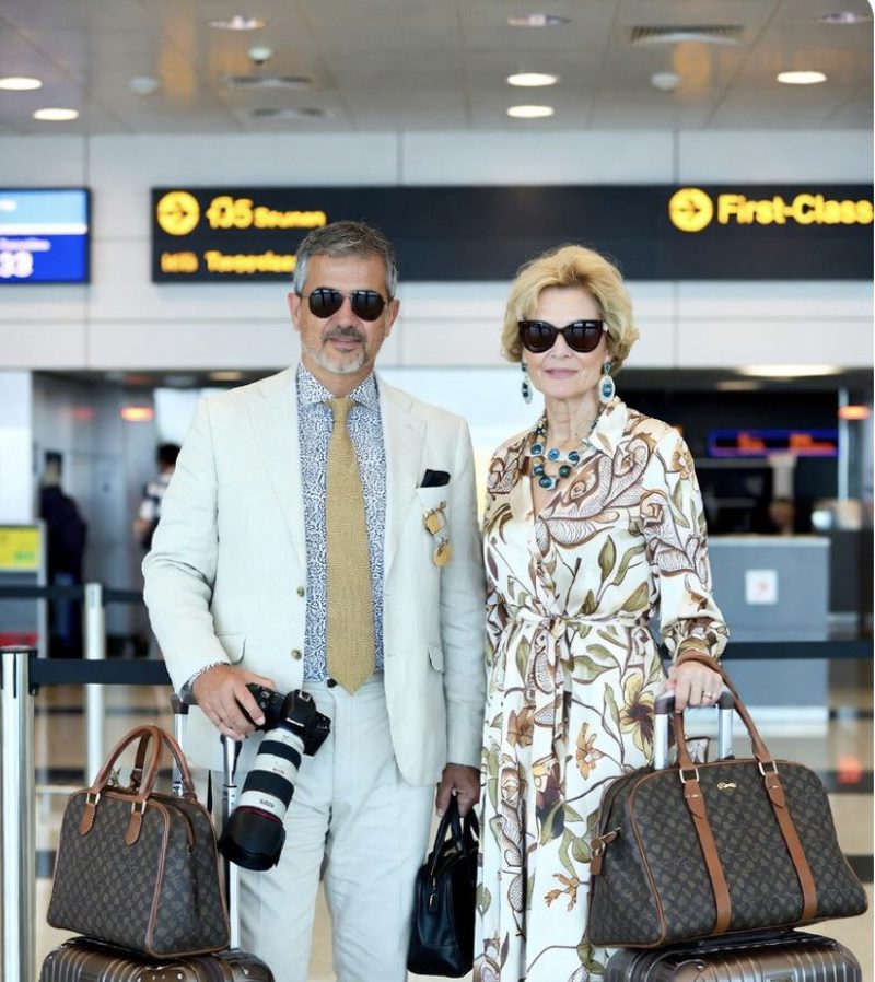 A well-dressed older couple in sunglasses with matching designer luggage and a large camera, standing under a First-Class airport sign