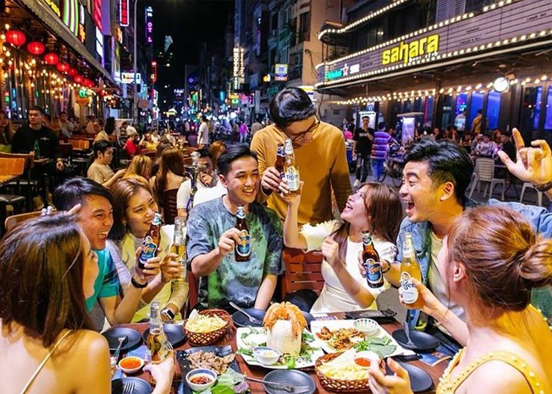 A lively night-time street-food table in Saigon, friends laughing and raising bottles